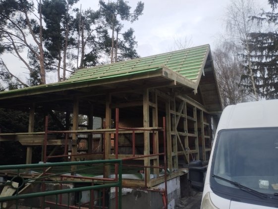 Konstrukcja drewniana z zielonym poszyciem dachu w trakcie budowy. Widoczne rusztowania i częściowo biały samochód dostawczy. Tło stanowią drzewa i pochmurne niebo.