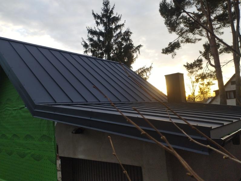 Nowoczesny, grafitowy dach z blachy na tle zachodzącego słońca i drzew. Widoczny komin i fragment ściany z zieloną membraną. Ujęcie z dołu, podkreślające geometrię dachu.