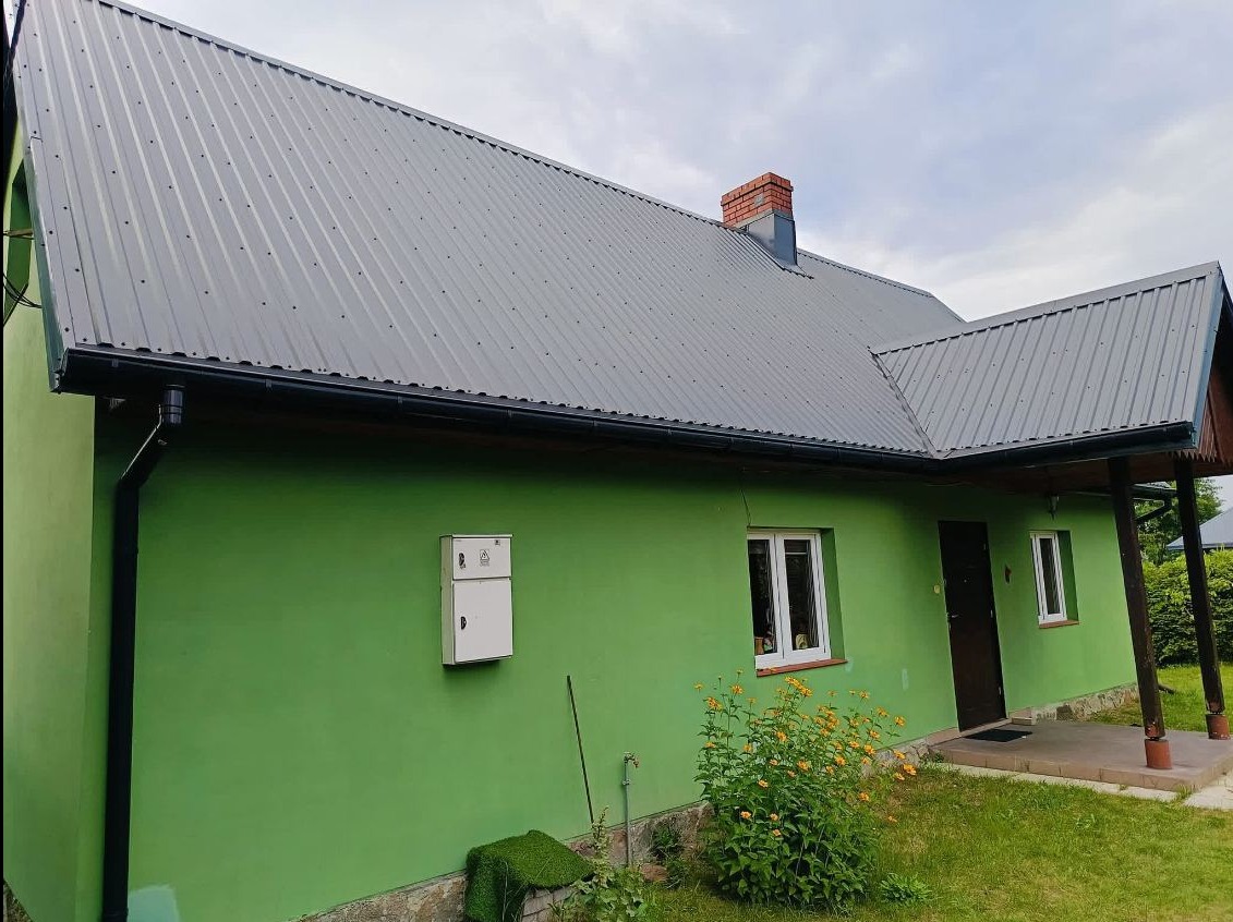 Szary dach z blachy trapezowej na zielonym domu jednorodzinnym w Chotomowie. Widoczne orynnowanie i komin z cegły. Ujęcie z poziomu gruntu.