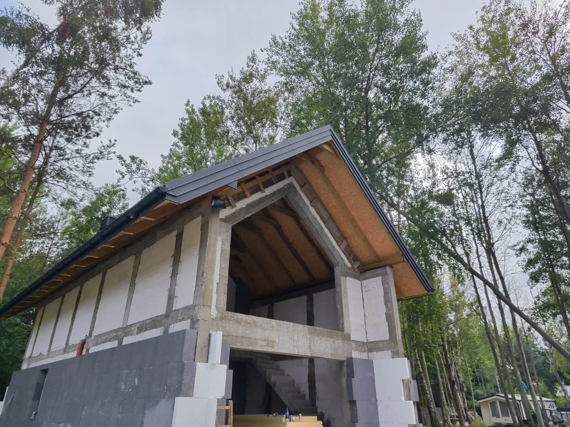 Budowa domu w lesie: widok na szkielet budynku z zamontowanym, czarnym dachem. Widoczne białe bloczki i drewniane elementy konstrukcyjne. W tle wysokie drzewa.