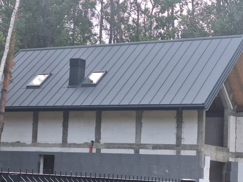 Szary dach z blachy na domu w trakcie budowy, widoczne okna dachowe i komin. Tło stanowią drzewa.