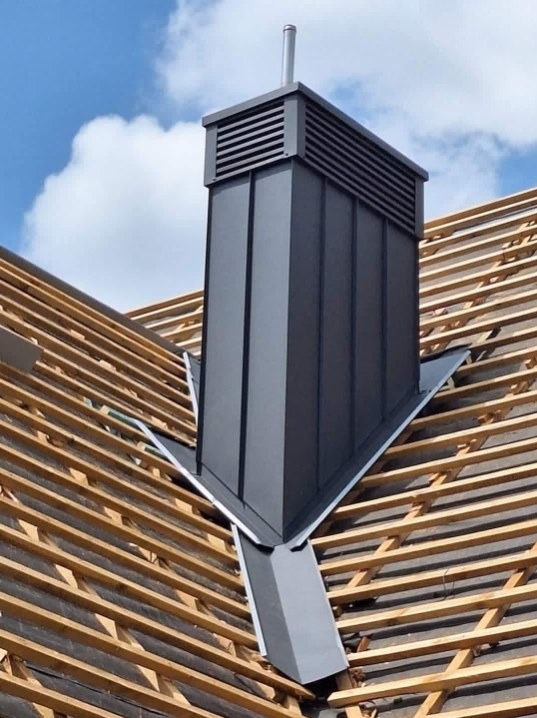 Nowoczesny, grafitowy komin wentylacyjny z blachy na tle drewnianej konstrukcji dachu i błękitnego nieba. Widoczne detale łączeń blacharskich.