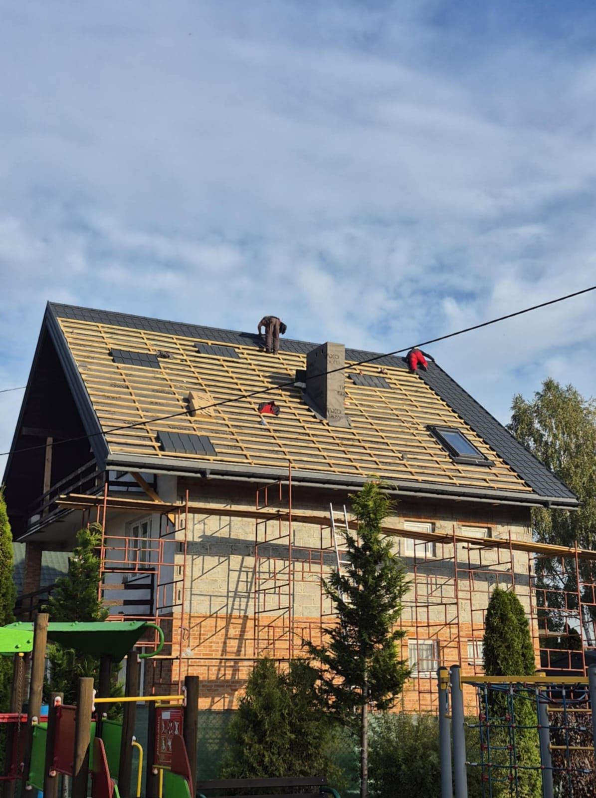 Dom w rusztowaniach z częściowo położonym, czarnym dachem. Dwóch dekarzy pracuje na dachu, układając dachówkę. Widoczne łatowanie i okno dachowe.