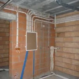 Surowy stan: ściany z cegły dziurawki, białe peszle z przewodami elektrycznymi ułożone równolegle pod sufitem i wzdłuż ściany, puszka elektryczna na ścianie, rury pod sufitem, betonowy sufit.