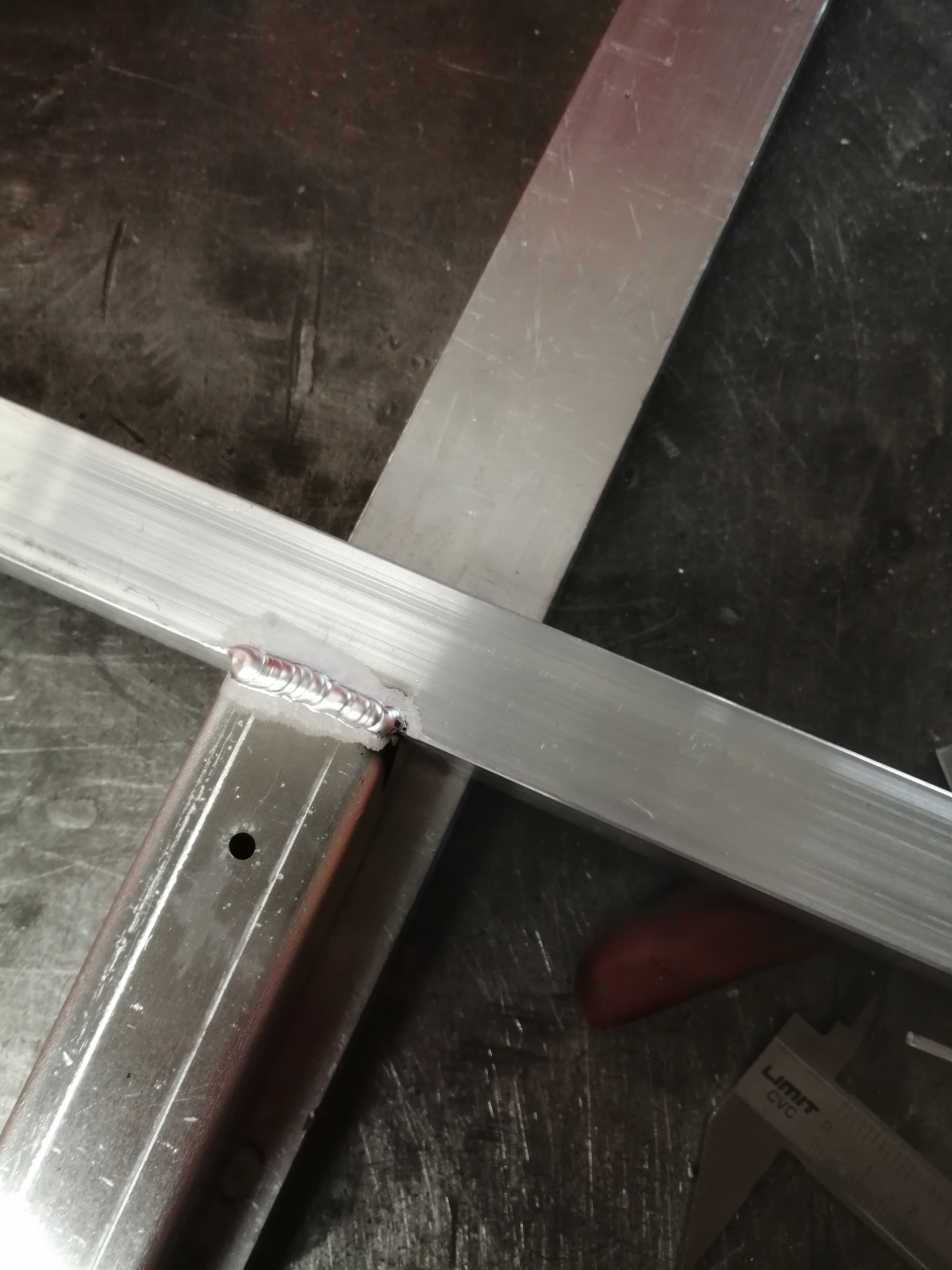 Spoina łącząca dwa aluminiowe profile, widoczne narzędzie pomiarowe i ślady obróbki na warsztatowym stole.