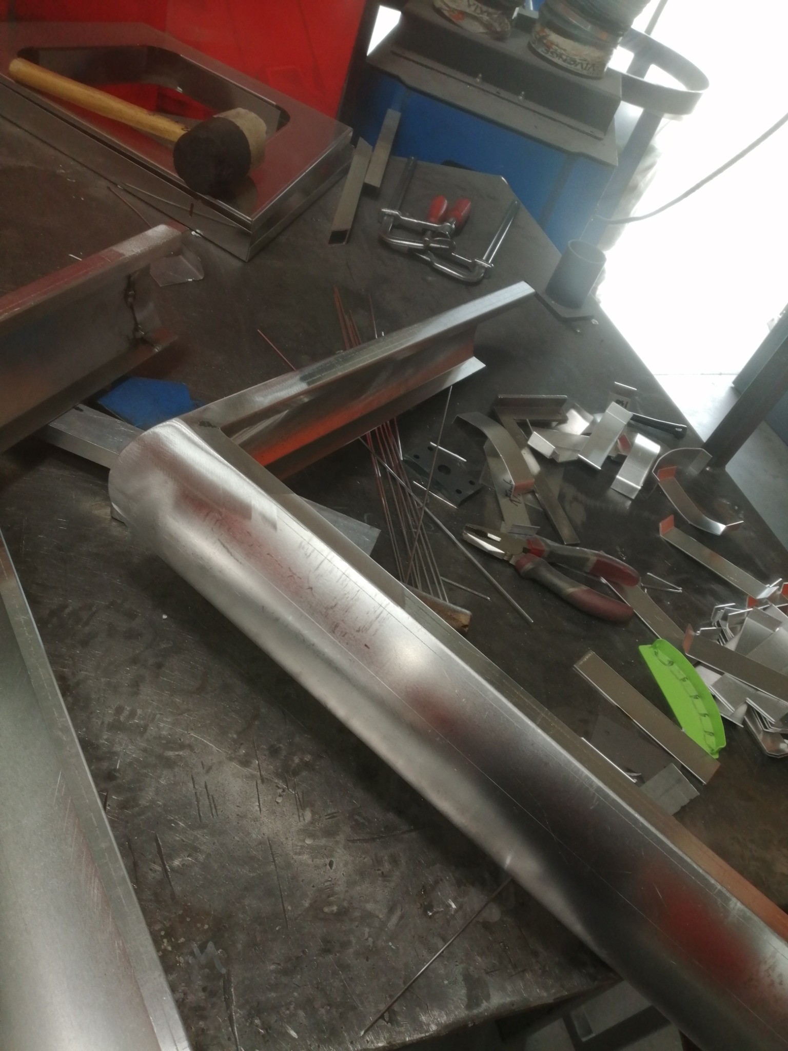 Warsztatowy stół roboczy z elementami metalowymi, narzędziami i gumowym młotkiem, ukazujący etap obróbki metalu.