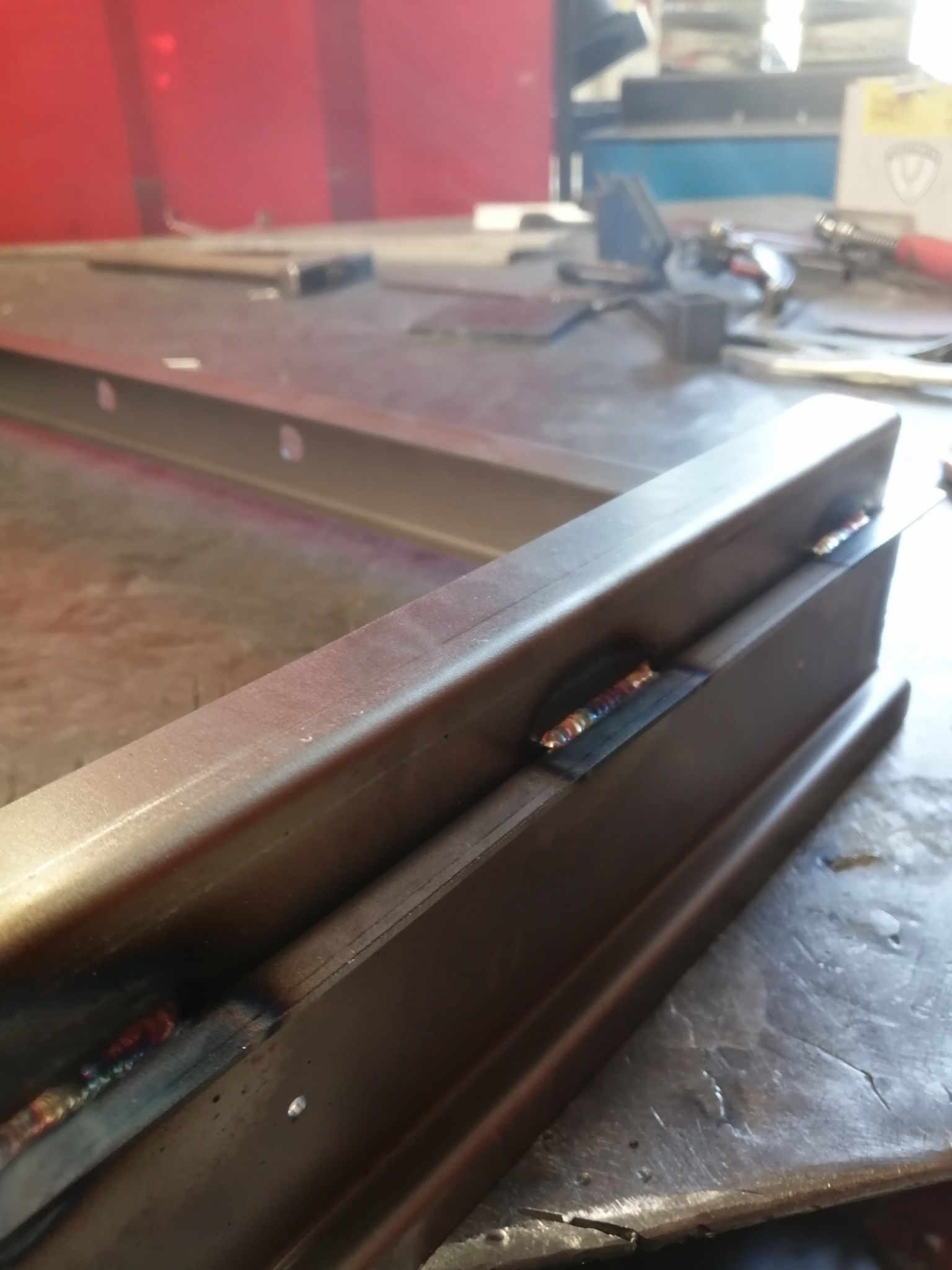 Spojenie spawane dwóch profili metalowych o przekroju kwadratowym, widoczne tęczowe przebarwienia na spoinie, surowy warsztatowy stół w tle.