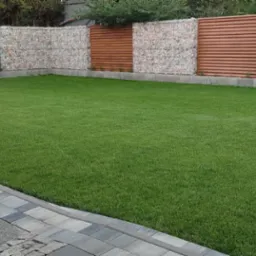 Starannie utrzymany trawnik z rolki, otoczony gabionami i drewnianym ogrodzeniem, z kostką brukową na obrzeżach, w słoneczny dzień.