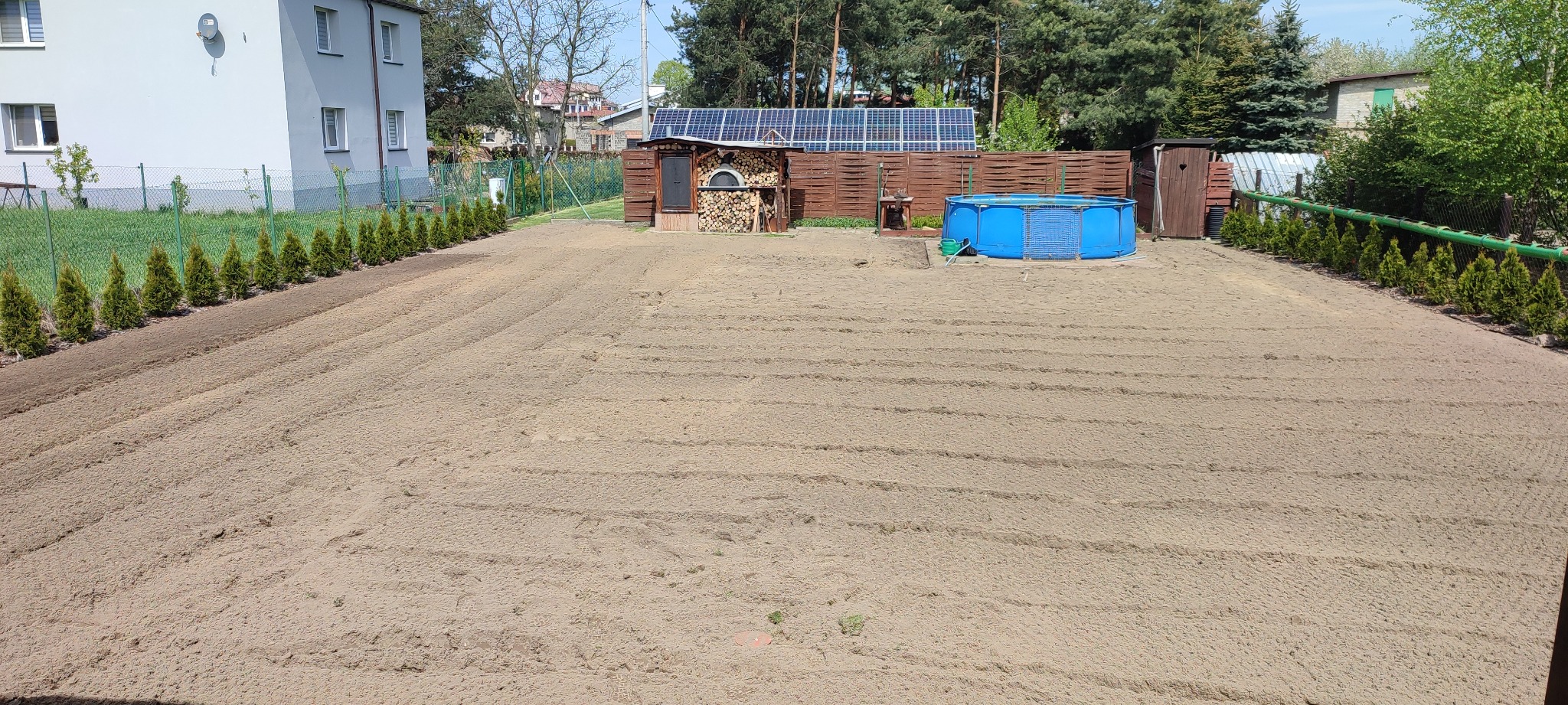 Przygotowany teren pod trawnik, z widocznymi śladami po bronowaniu, ogrodzony tujami, z basenem ogrodowym w tle i budynkiem gospodarczym z panelami słonecznymi na dachu.