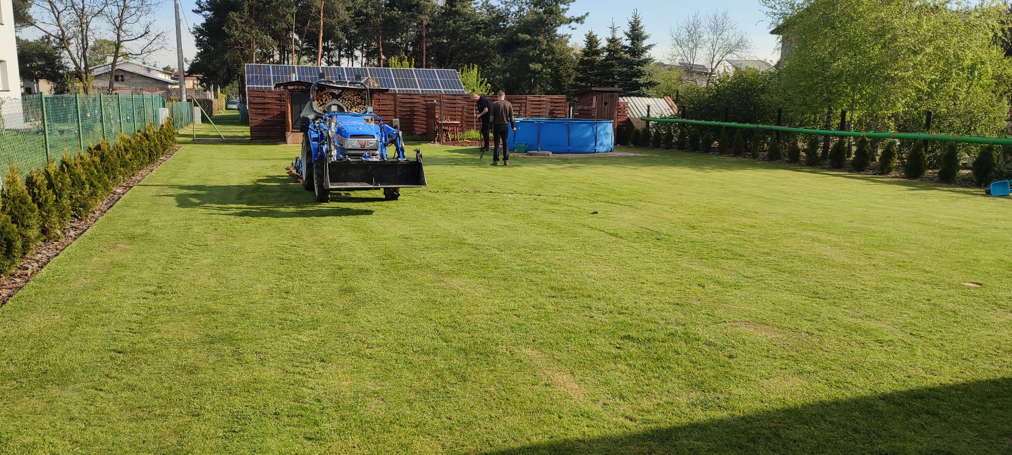 Niebieski traktorek z łopatą spulchnia ziemię na trawniku, w tle pracownicy oraz basen ogrodowy i zabudowania z panelami solarnymi.