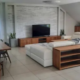 Jasny salon z narożną sofą z białej skóry, drewnianym stolikiem kawowym z półkami, drewnianą szafką RTV pod telewizorem i drewnianym regałem w zabudowie pod skosem dachu.