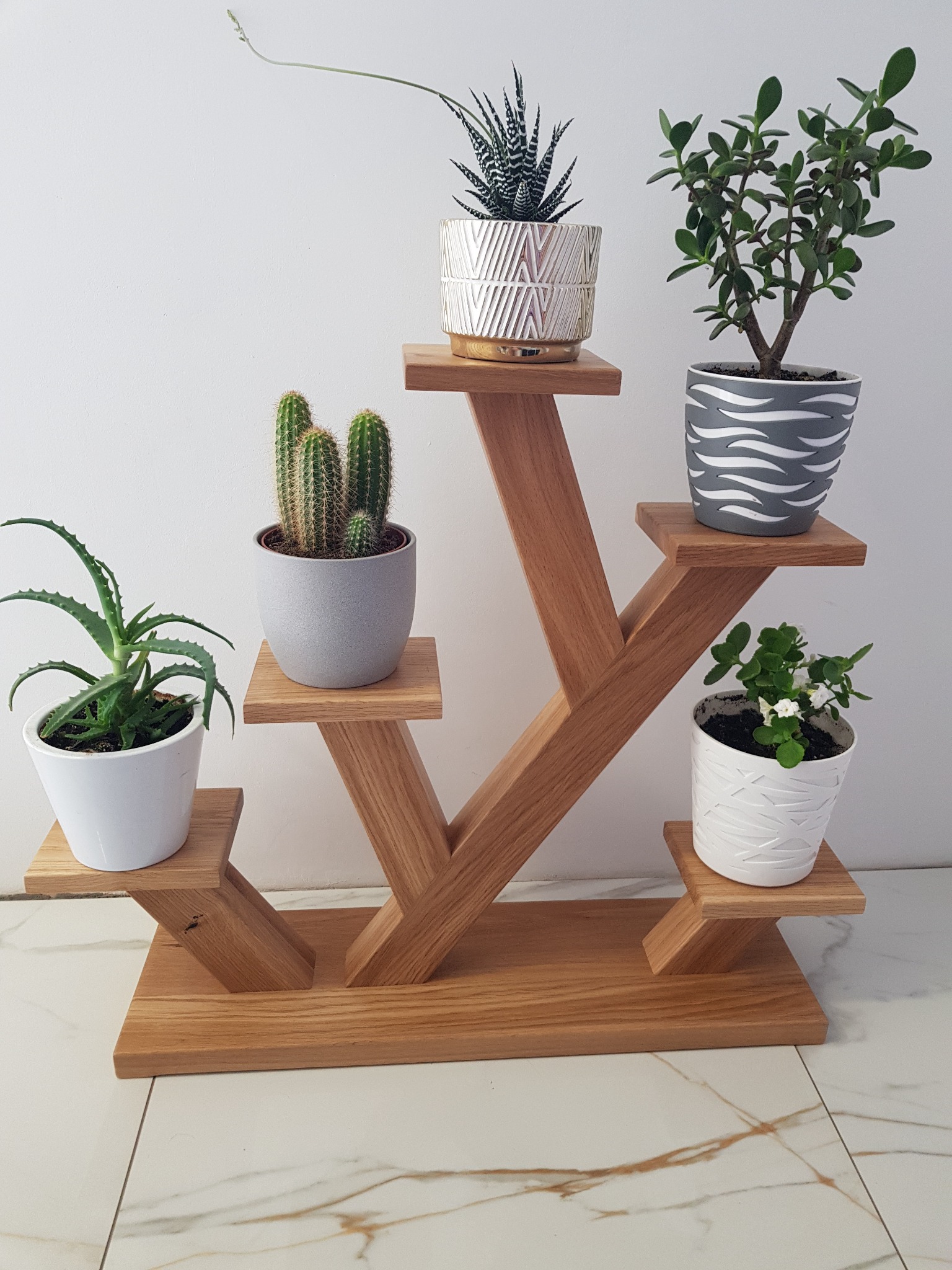 Drewniany regał na rośliny z pięcioma półkami, na których stoją doniczki z różnymi kwiatami, na tle białej ściany i marmurowej podłogi.