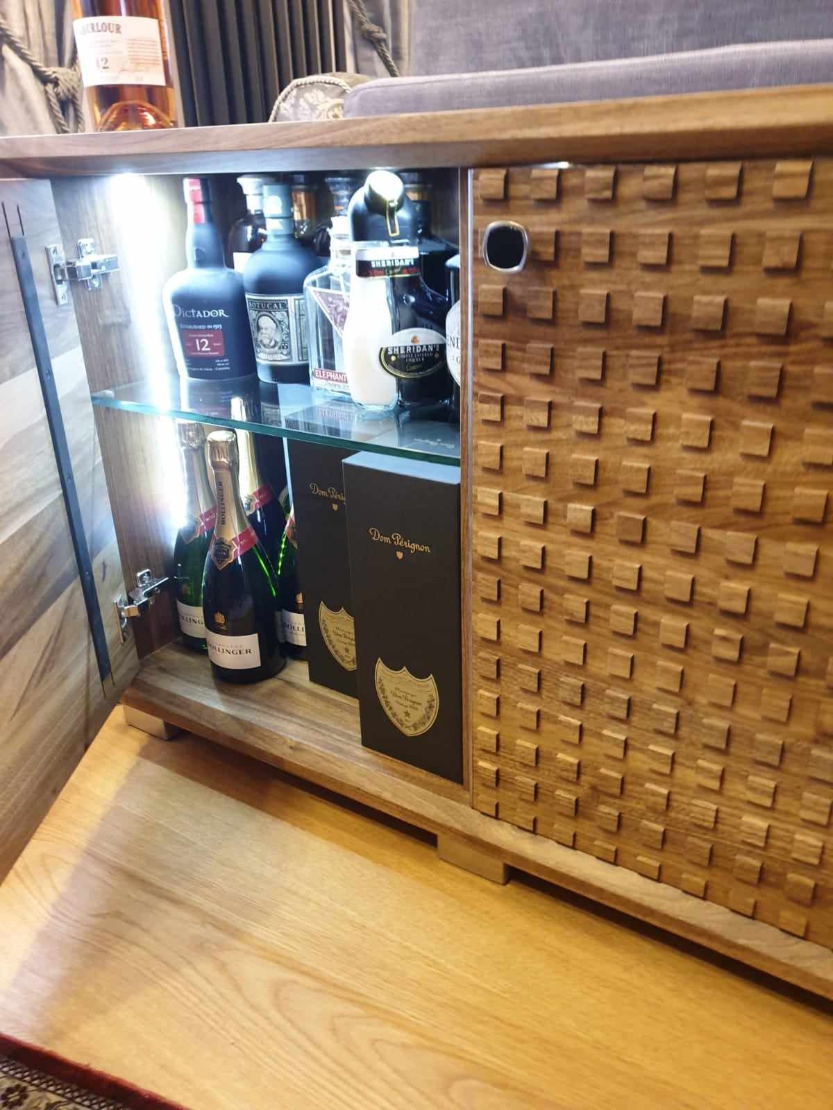 Drewniana szafka barowa z oświetleniem LED, półkami z przezroczystego szkła i dekoracyjną frontową ścianką z wystającymi kwadratami. Wewnątrz widoczne butelki alkoholi i szampana, w tym Dom Pérignon.