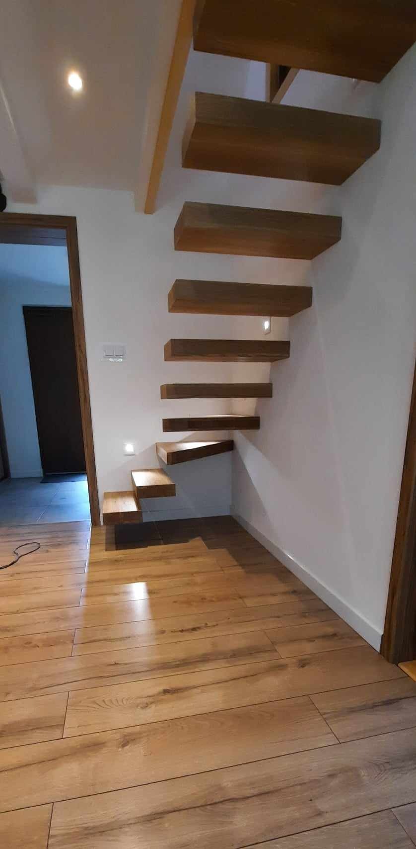 Drewniane schody wspornikowe zamontowane przy ścianie z oświetleniem punktowym, widoczne drewniane wykończenie podłogi i drzwi.