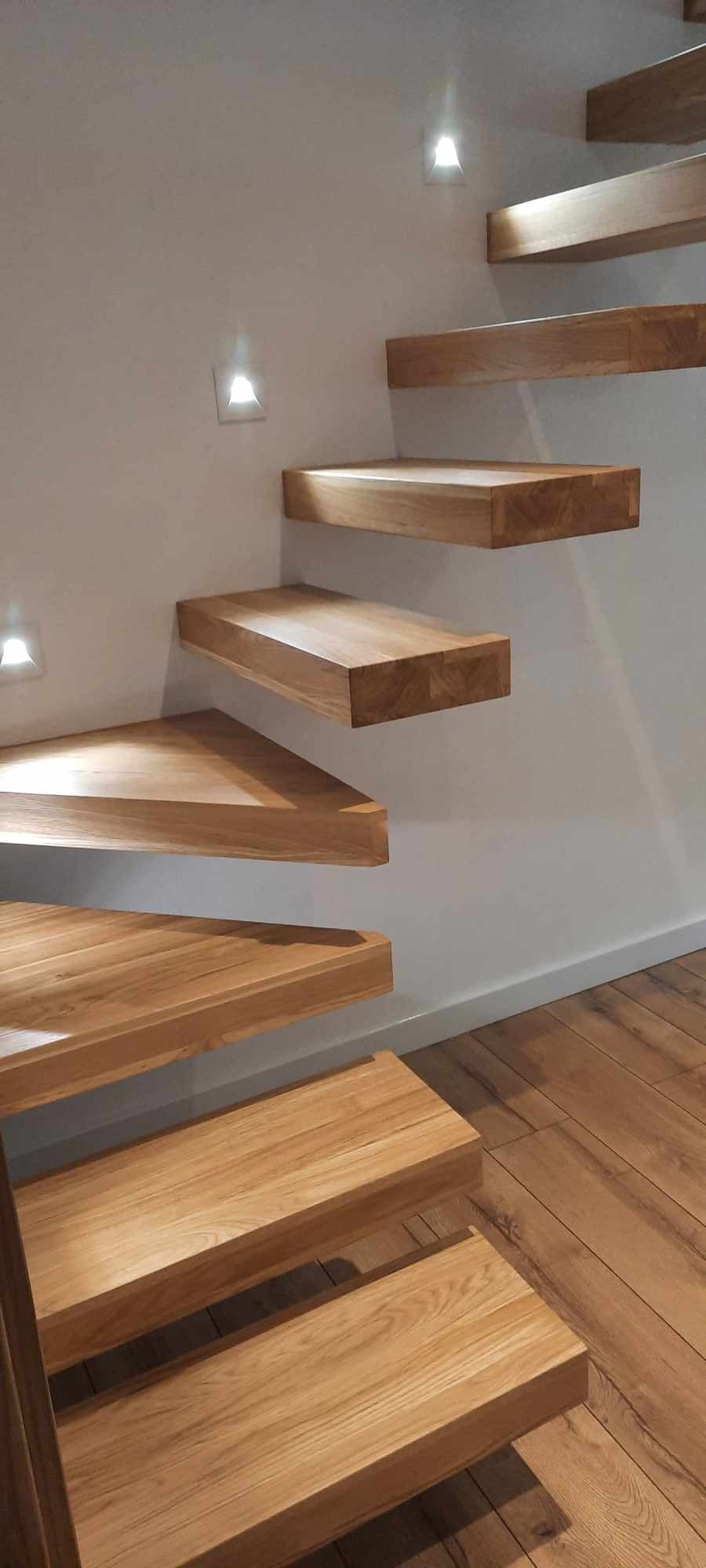 Drewniane schody wiszące o prostej formie, oświetlone punktowymi lampami w ścianie, z parkietem na podłodze.