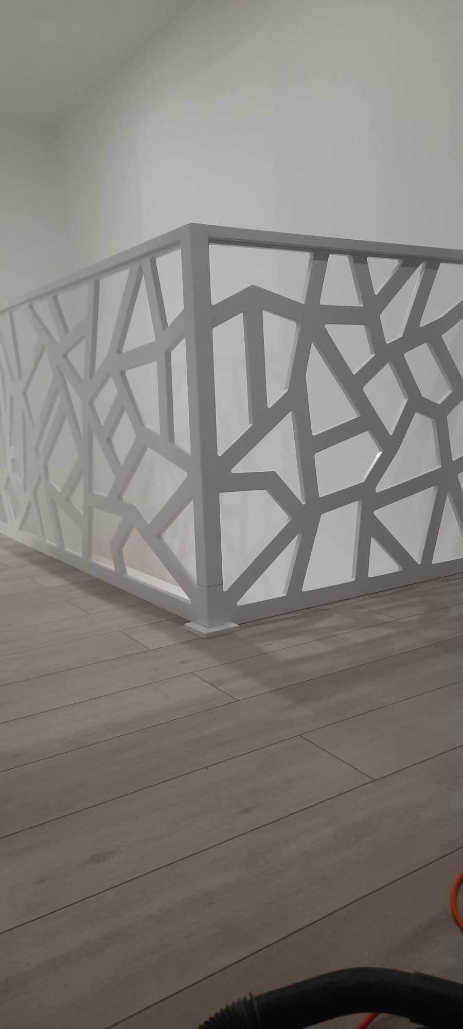Biała balustrada schodowa z geometrycznym ażurem, zamontowana na drewnianej podłodze, minimalistyczny design.