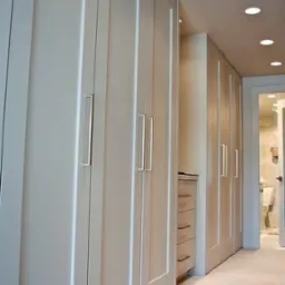 Biała szafa wnękowa z długimi srebrnymi uchwytami, widoczna z perspektywy korytarza z otwartymi drzwiami do łazienki.