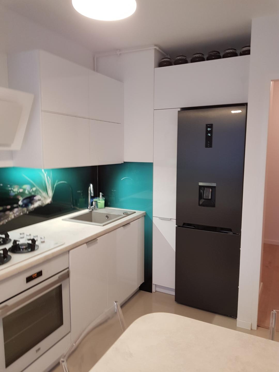 Nowoczesna kuchnia z białymi, lakierowanymi frontami, turkusowym panelem szklanym nad blatem i czarną lodówką z dozownikiem wody.