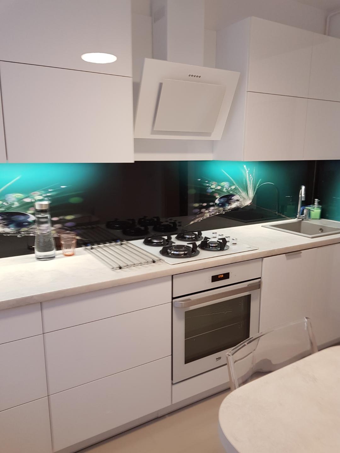 Minimalistyczna biała kuchnia z błyszczącym panelem szklanym z motywem dmuchawca, płyta gazowa i piekarnik w zabudowie.