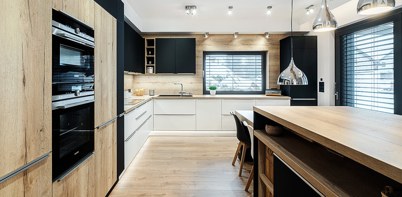 Nowoczesna kuchnia z drewnianymi i czarnymi szafkami, białymi blatami, podświetleniem LED pod szafkami i wyspą kuchenną z drewnianym blatem.