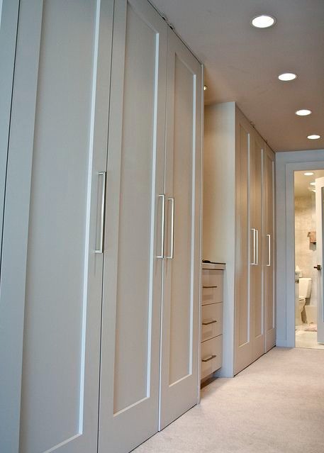 Biała szafa wnękowa z długimi srebrnymi uchwytami, widoczna z perspektywy korytarza z otwartymi drzwiami do łazienki.