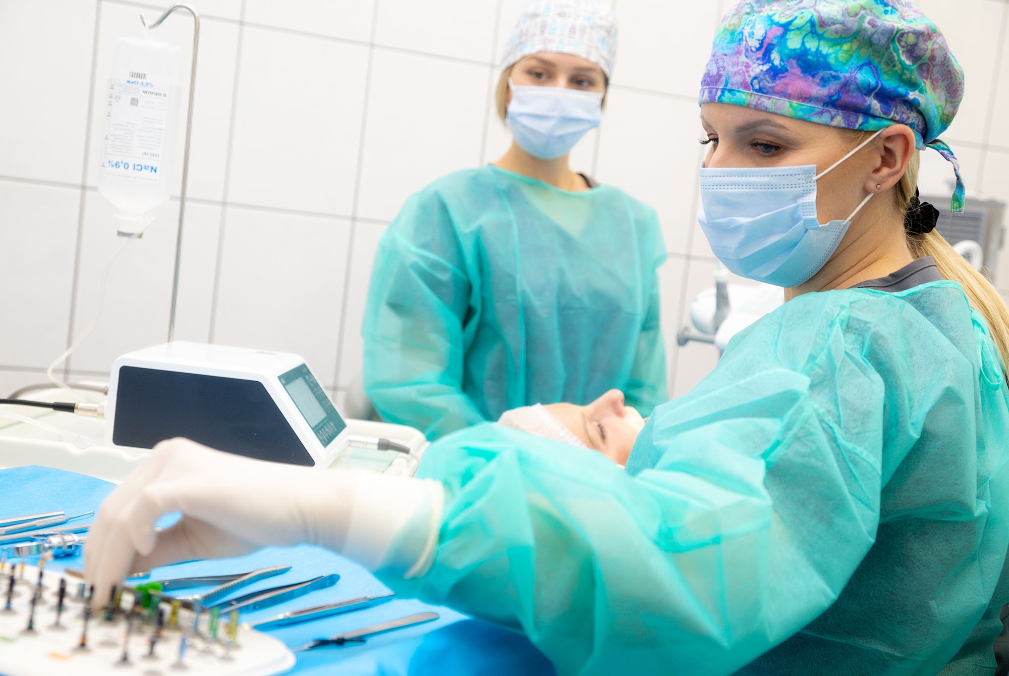 Stomatolog w trakcie zabiegu, asystentka w tle, narzędzia stomatologiczne na stole, kroplówka obok, białe kafelki na ścianie.