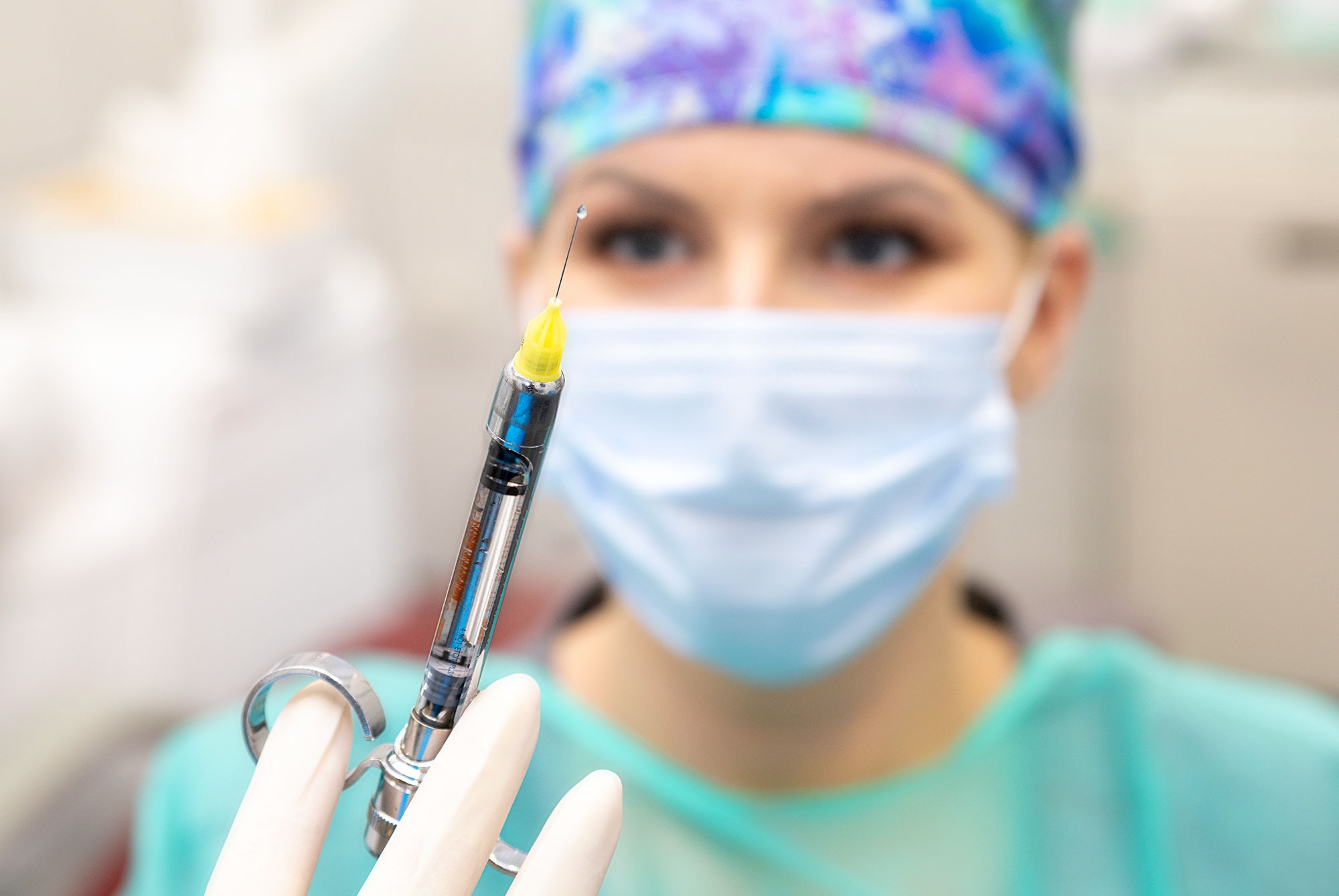 Lekarz w masce i czepku trzyma w rękawicach strzykawkę z kroplą na końcu igły, przygotowując się do zabiegu.