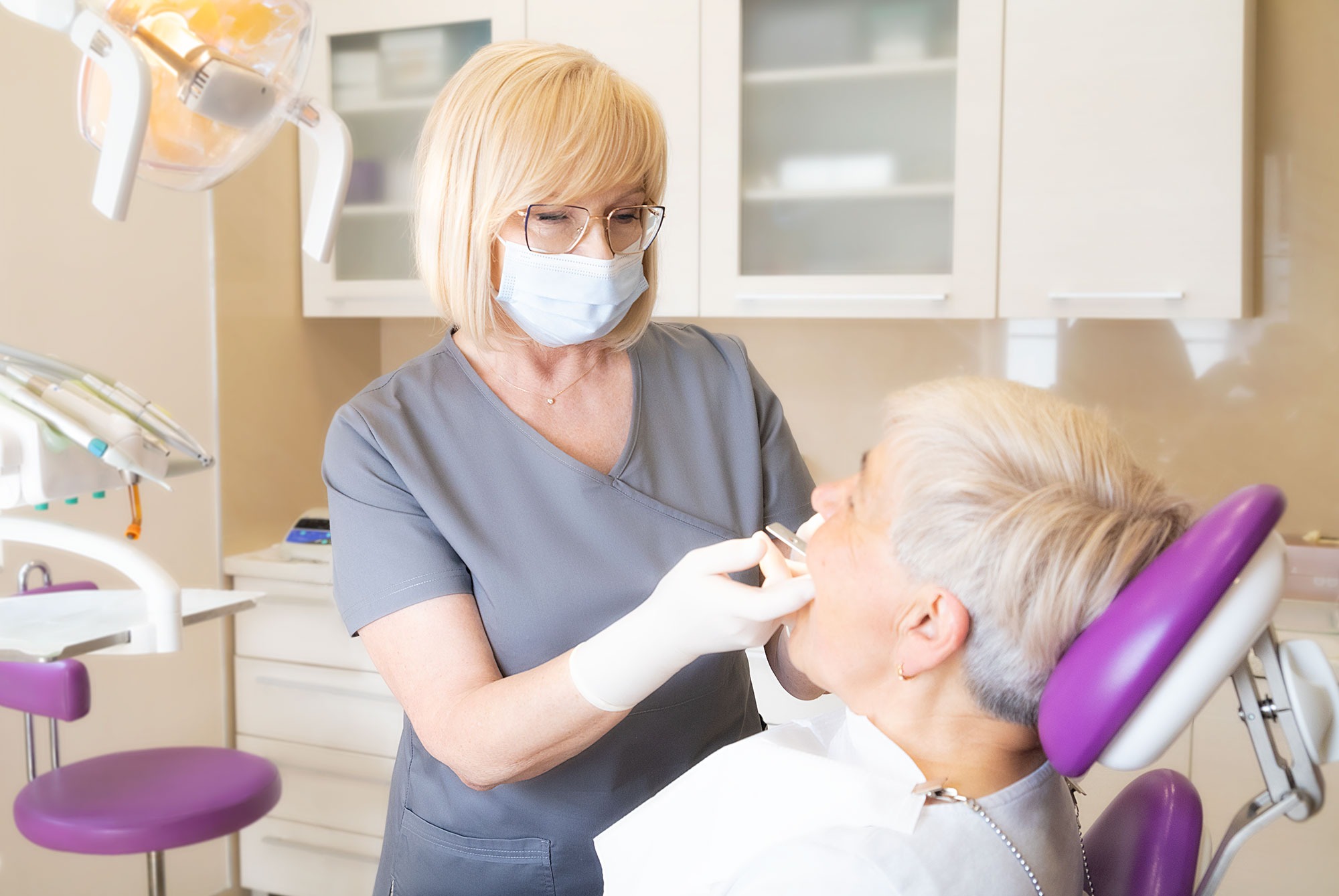 Lekarz w okularach i maseczce, w trakcie badania jamy ustnej pacjentki w fotelu dentystycznym, widoczna lampa zabiegowa i meble medyczne.
