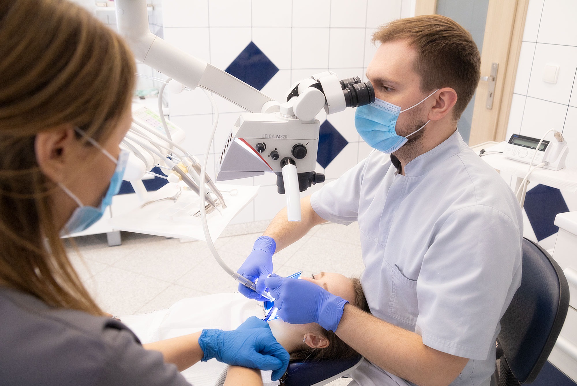 Lekarz w masce i rękawicach, używający mikroskopu Leica M320 podczas zabiegu na pacjencie leżącym na fotelu, asystuje druga osoba.