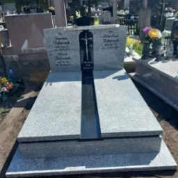Granitowy nagrobek podwójny z czarnym pasem pośrodku, na cmentarzu, z wygrawerowanymi imionami i datami.