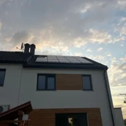 Biały dom z panelami słonecznymi na ciemnym dachu, widoczne kominy i antena satelitarna, na tle pochmurnego nieba o zmierzchu. Częściowe drewniane wykończenie elewacji.