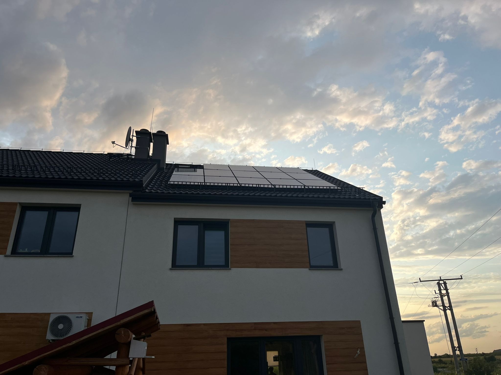 Biały dom z panelami słonecznymi na ciemnym dachu, widoczne kominy i antena satelitarna, na tle pochmurnego nieba o zmierzchu. Częściowe drewniane wykończenie elewacji.
