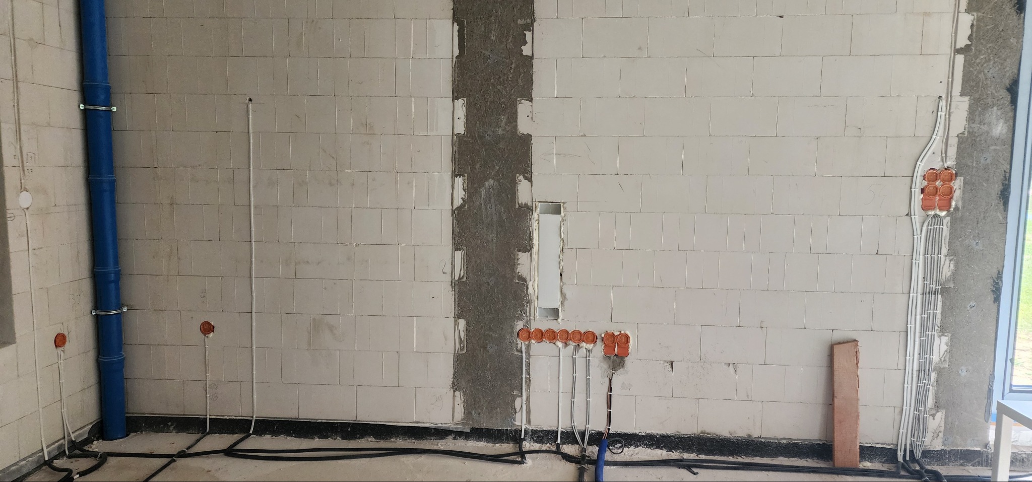Instalacja elektryczna w trakcie montażu: kable w ścianie z bloczków, puszki instalacyjne, rury. Widoczne elementy instalacji wodnej. Surowy stan wykończenia wnętrza.