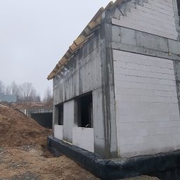 R.B Remonty - Surowy stan budynku w trakcie budowy, widoczne białe bloczki, szara konstrukcja betonowa, drewniane elementy dachu i czarna folia izolacyjna przy fundamencie.