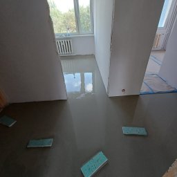 R.B Remonty - Wylewka samopoziomująca w trakcie realizacji. Widoczne fragmenty ścian, okno z widokiem na zieleń i poziomice. Świeża, mokra powierzchnia wylewki odbija światło.