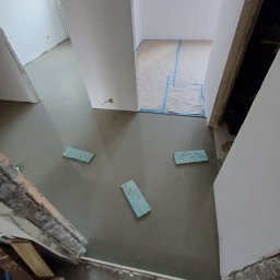 R.B Remonty - Wylewka samopoziomująca w trakcie prac, widoczne fragmenty pianki dylatacyjnej i przygotowane pomieszczenie z oknem. Ujęcie z góry, surowy stan.