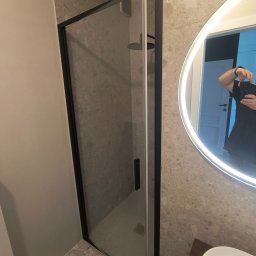 R.B Remonty - Minimalistyczna łazienka z przeszkloną kabiną prysznicową z czarnym profilem, podświetlanym okrągłym lustrem i ścianami wyłożonymi płytkami imitującymi lastryko.