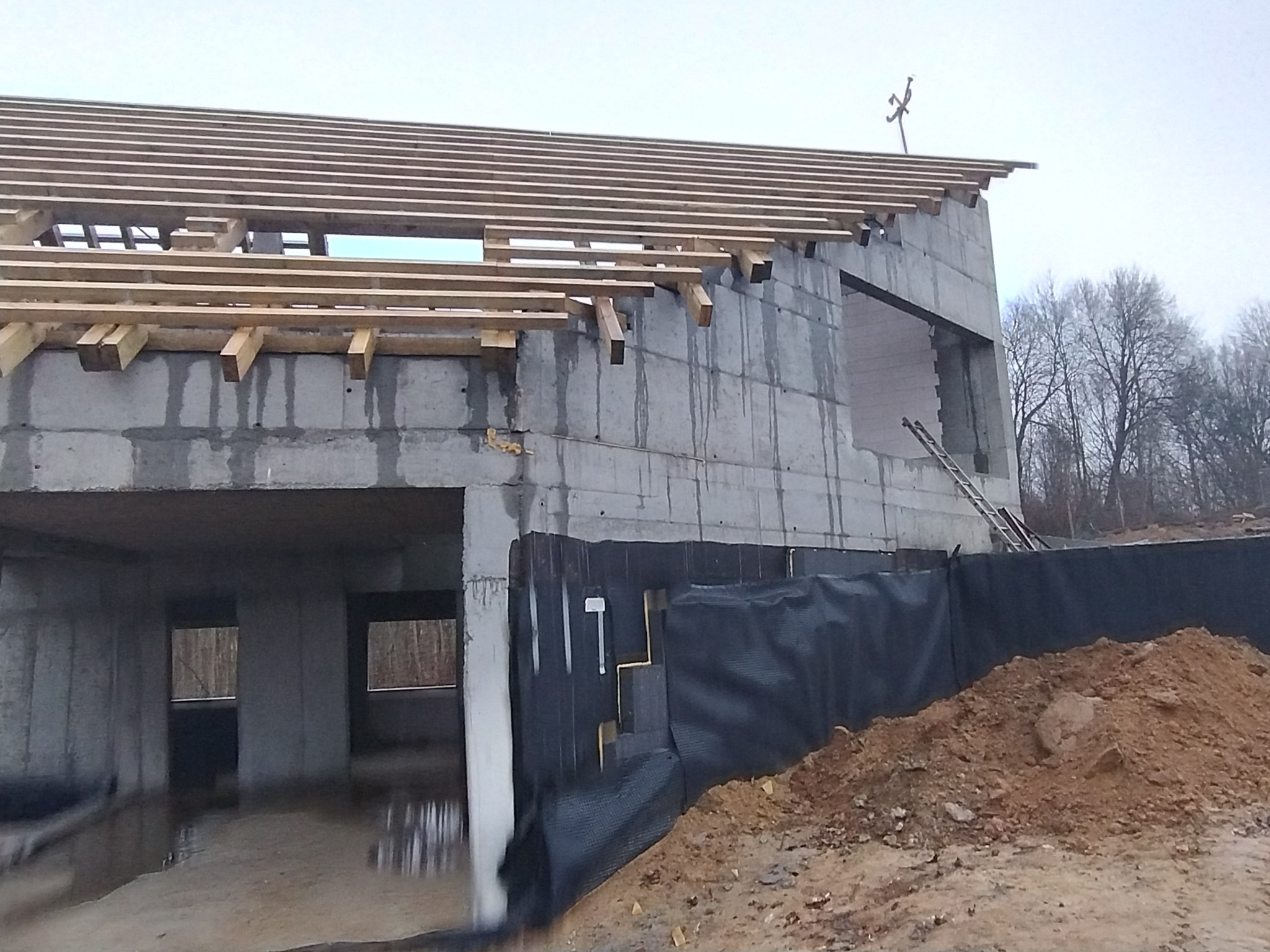 Surowy stan budynku z betonu komórkowego w trakcie budowy, widoczne drewniane elementy konstrukcji dachu, czarna folia izolacyjna i sterta ziemi.