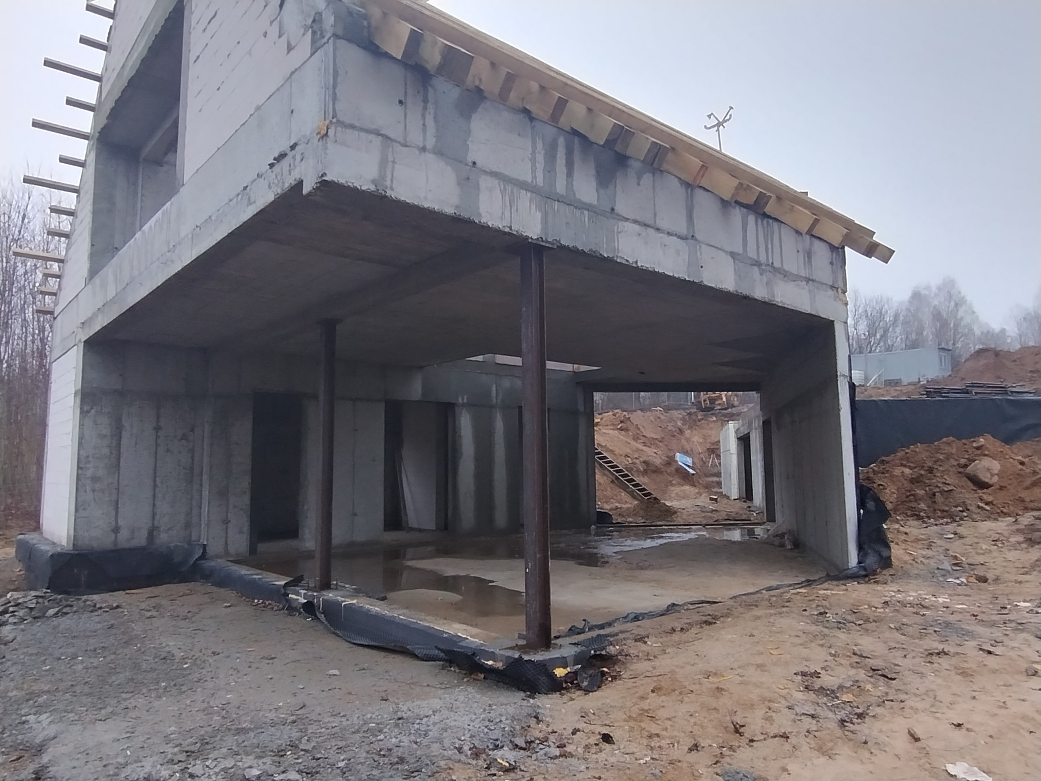Surowy stan budynku w trakcie budowy, widoczne betonowe ściany z pustaków, stalowe podpory, drewniane elementy konstrukcji dachu oraz teren wokół budynku w trakcie prac ziemnych.