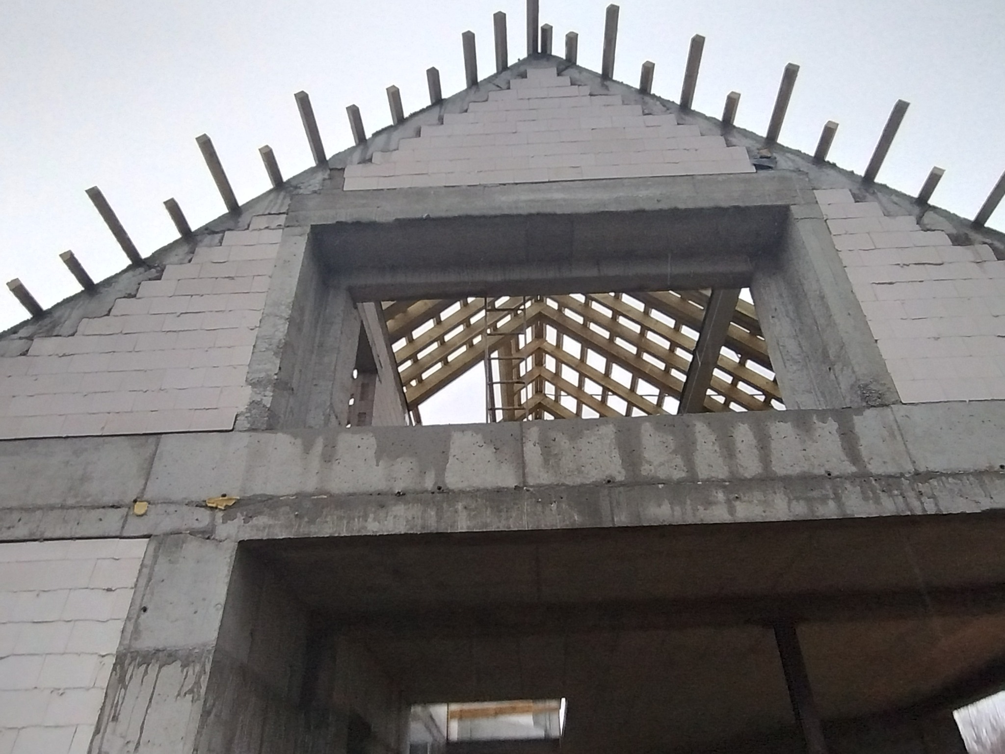 Surowy stan budynku w trakcie budowy: ściany z betonu komórkowego, drewniana konstrukcja dachu widoczna przez otwór okienny, wystające drewniane elementy na szczycie dachu.