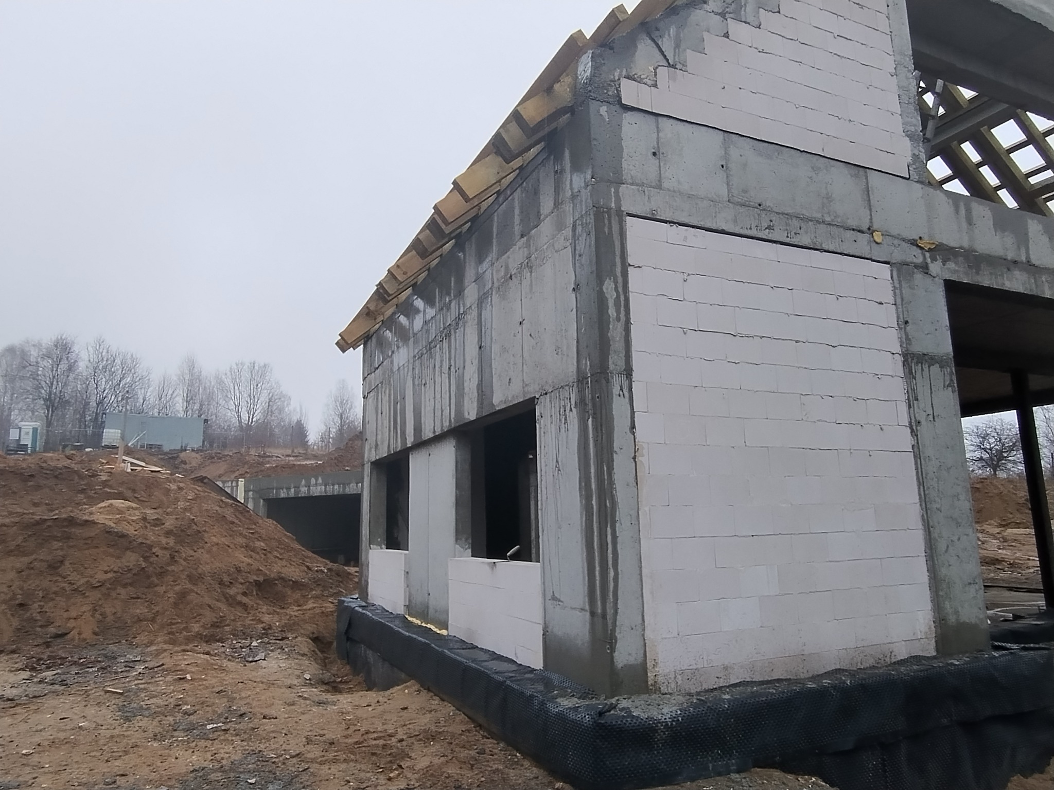 Surowy stan budynku w trakcie budowy, widoczne białe bloczki, szara konstrukcja betonowa, drewniane elementy dachu i czarna folia izolacyjna przy fundamencie.