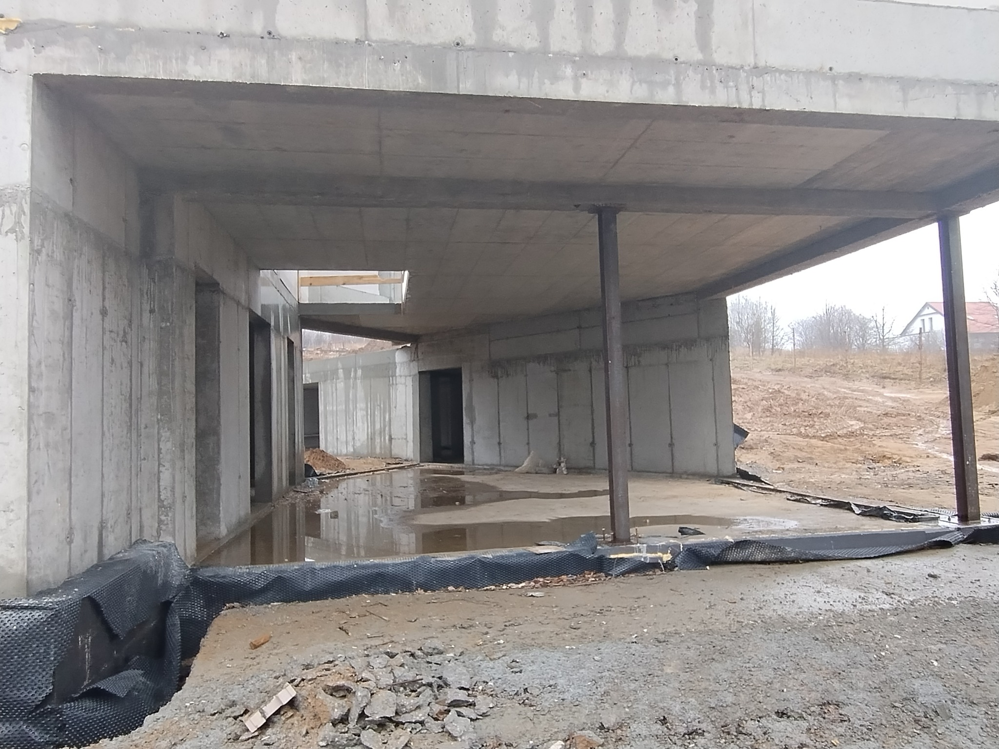 Surowy betonowy szkielet budynku w trakcie budowy, widoczne wsporniki i izolacja fundamentów, ziemia i kałuże na podłożu.