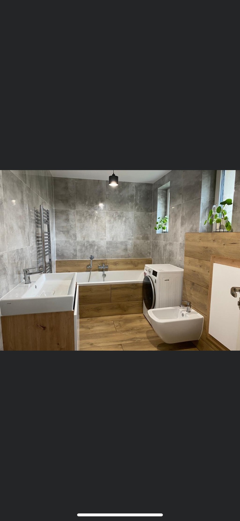 Nowoczesna łazienka z szarymi płytkami, drewnopodobną zabudową wanny i umywalki, białą pralką i bidetem. Dwie roślinki w oknach dodają świeżości.