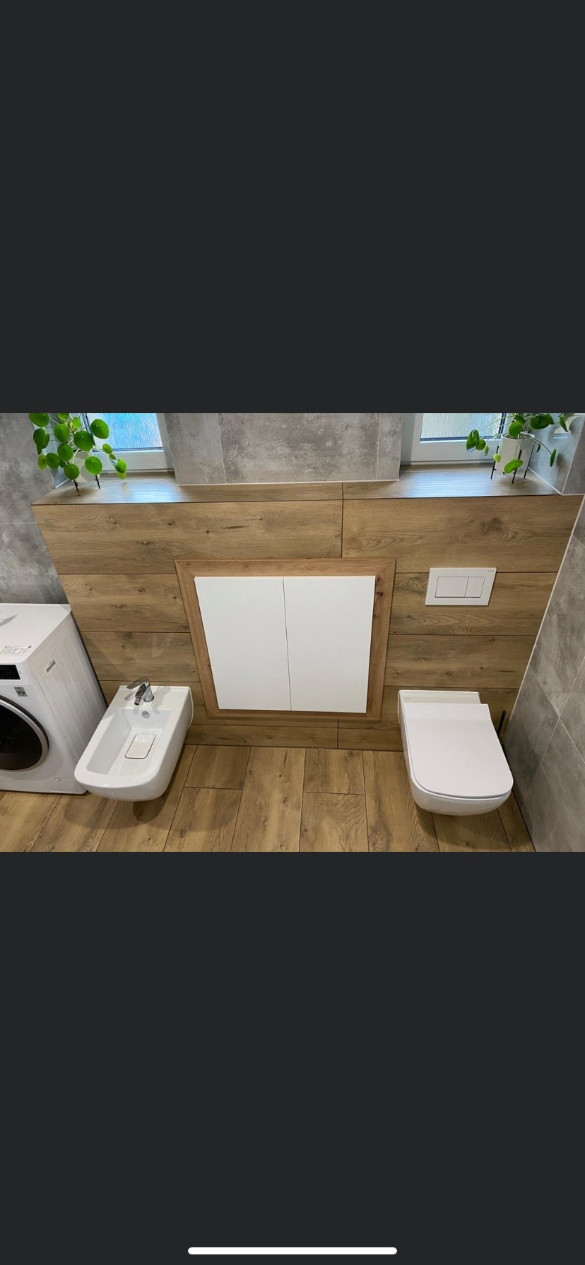 Nowoczesna łazienka z drewnianymi panelami i ukrytą szafką, wiszący bidet i toaleta, pralka z lewej strony, roślina doniczkowa na parapecie.