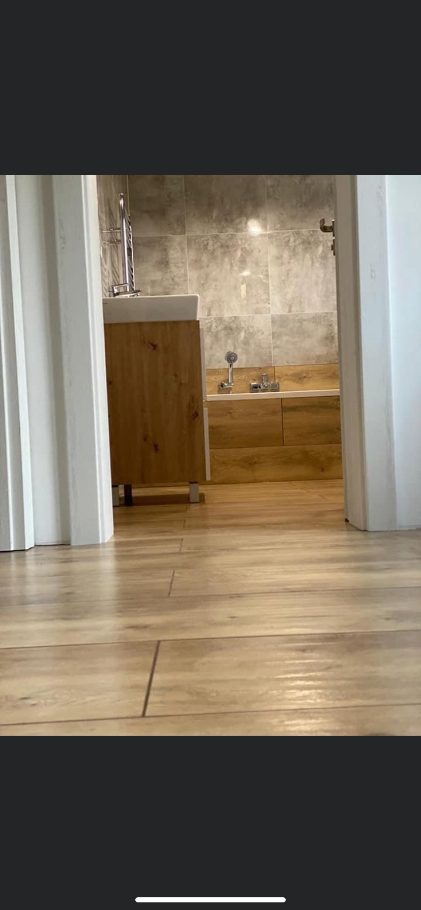 Widok z poziomu podłogi na wykończoną łazienkę z drewnopodobną zabudową wanny, umywalką na szafce i szarą glazurą na ścianach, widzianą przez otwarte drzwi.