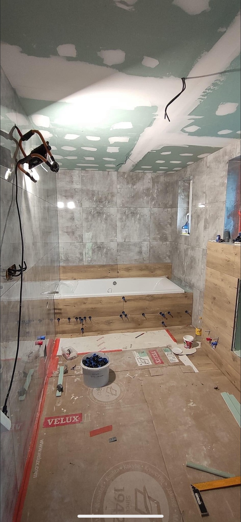 Łazienka w trakcie wykańczania: szare płytki na ścianach, obudowa wanny z drewnopodobnych paneli, sufit z płyt kartonowo-gipsowych przed malowaniem, widoczne narzędzia i akcesoria glazurnicze.