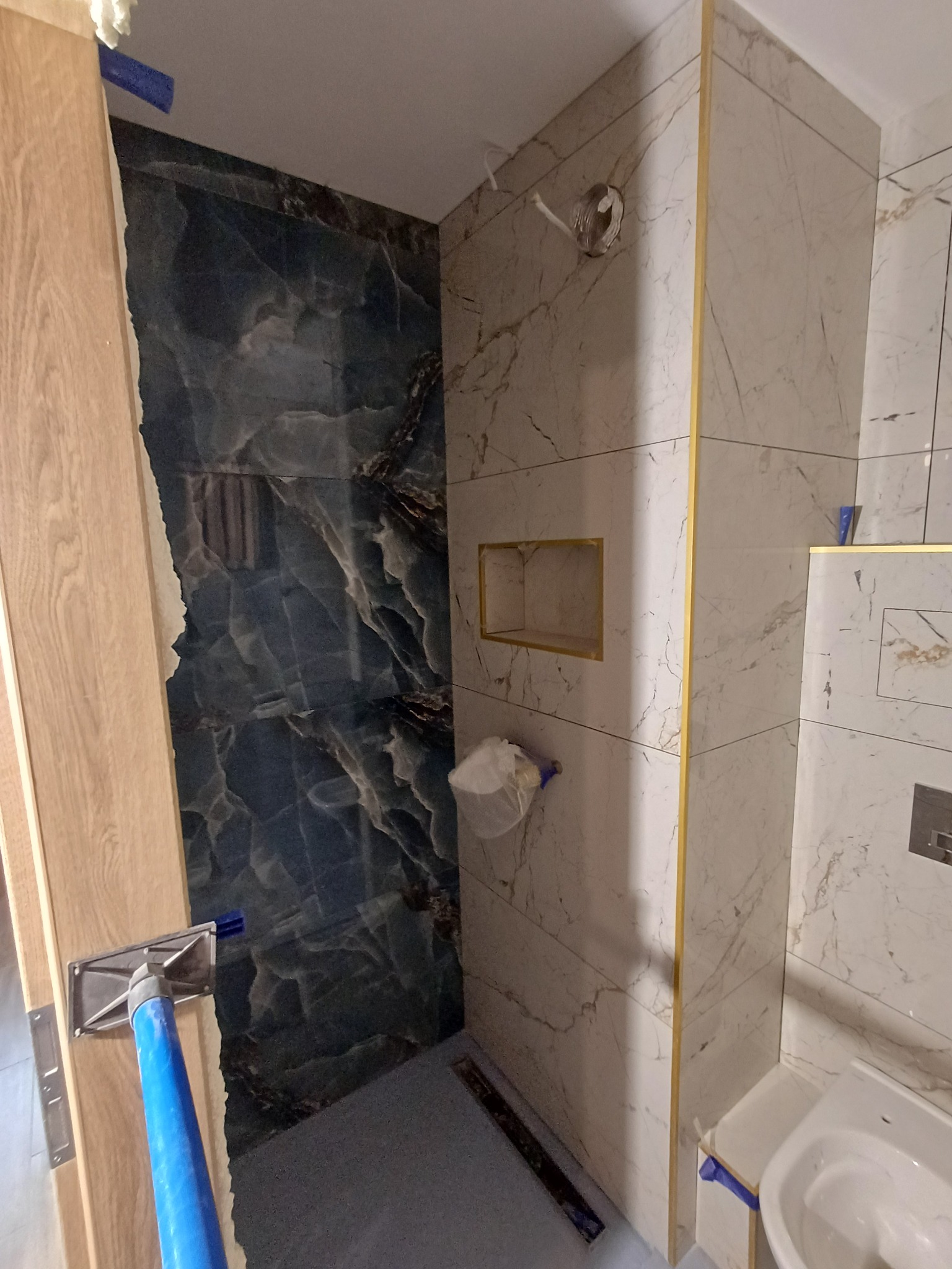 Fragment łazienki w trakcie wykańczania: położone marmurowe płytki w odcieniach bieli i granatu, widoczna wnęka w ścianie i narzędzie glazurnicze przy futrynie drzwi.