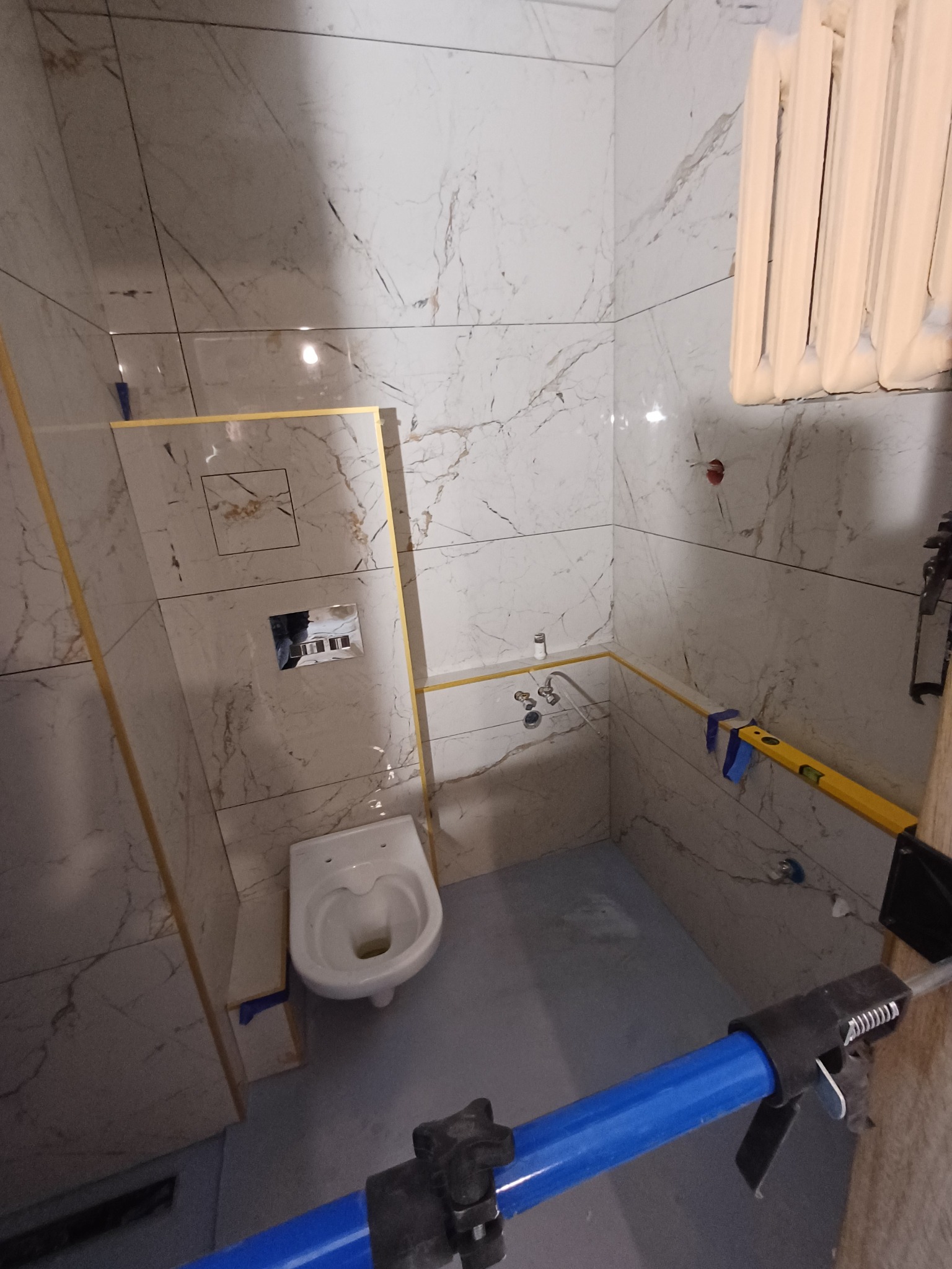 Łazienka w trakcie wykończenia: marmurowe płytki na ścianach, zamontowana toaleta, poziomica i narzędzia glazurnicze. Widoczne elementy instalacji i grzejnik.