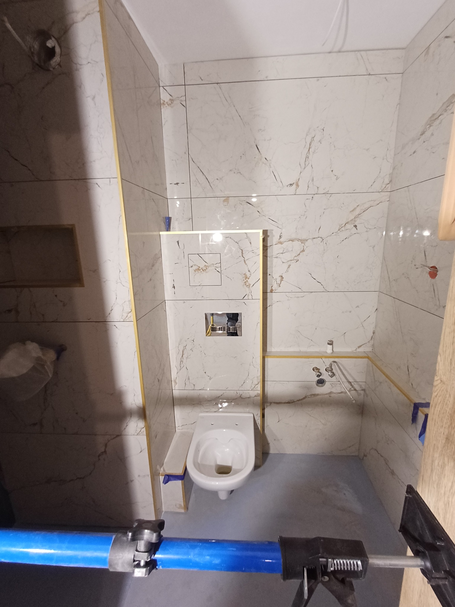 Biała łazienka z marmurowymi płytkami, widok na toaletę i fragment niebieskiego narzędzia, złote listwy wykończeniowe, surowy stan podłogi.