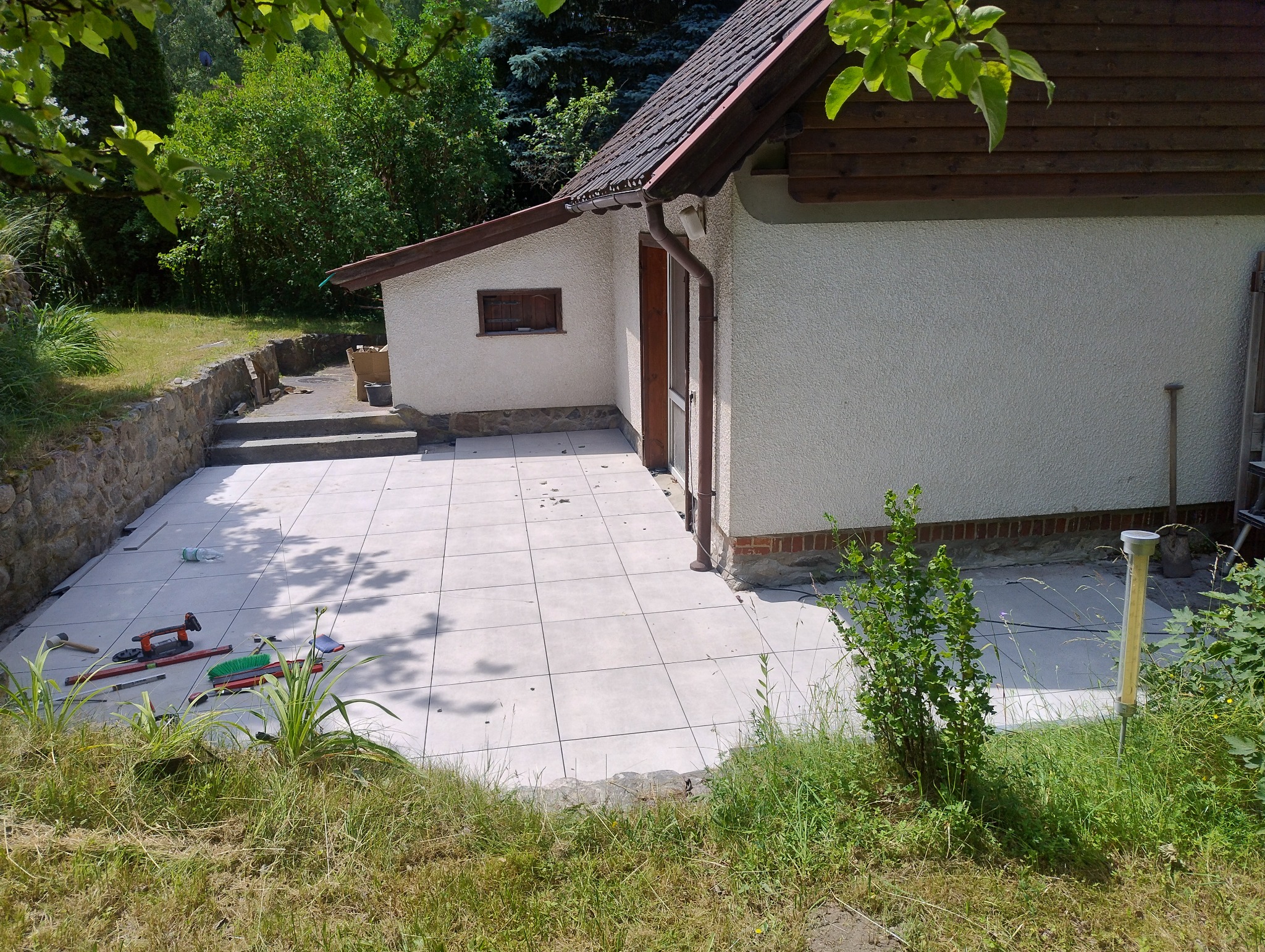 Jasne płytki tarasowe ułożone przy budynku z drewnianymi elementami. Widoczne narzędzia glazurnicze i zieleń w tle. Ukończony fragment tarasu.